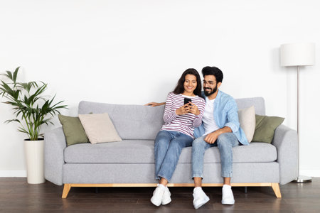 Relaxed multicultural couple sitting on sofa, using smartphoneの写真素材
