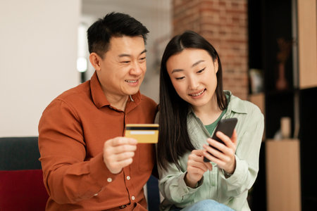 Cheerful asian husband and wife shopping online with smartphone and credit card, shopaholics enjoy sale togetherの写真素材