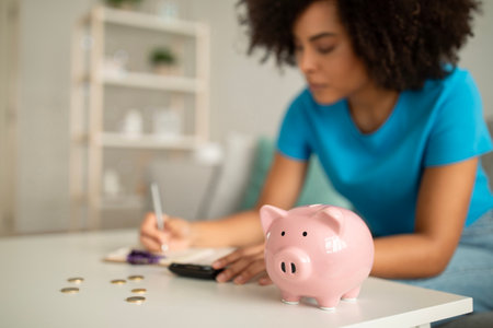 Serious concentrated millennial african american curly lady counting money near piggy bank in living roomの写真素材