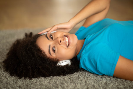 Glad millennial african american curly female in wireless headphones listen music, lying on floor, enjoy spare timeの写真素材
