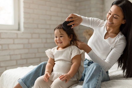 Happy Asian Mother Brushing Baby Daughters Hair Sitting In Bedroomの写真素材