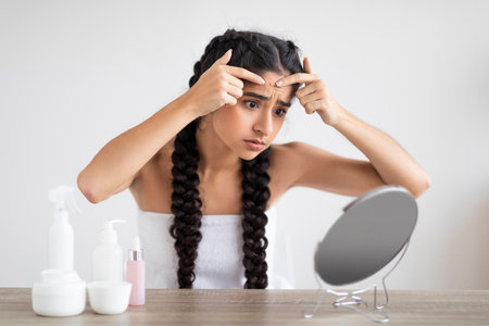 Upset Young Indian Female Popping Pimple On Forehead While Looking In Mirrorの写真素材