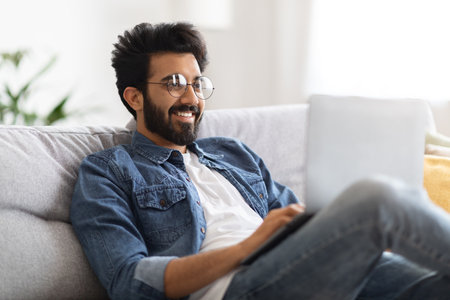 Smiling Handsome Indian Man Using Laptop While Sitting On Couch At Homeの写真素材