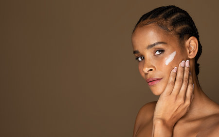 Portrait of black middle aged woman applying anti-aging or moisturising face cream, brown background, copy spaceの写真素材