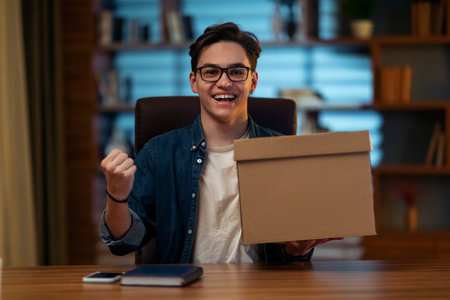 Satisfied young man sitting at workdesk, holding delivery box, gesturingの写真素材