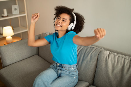 Glad millennial african american curly female in wireless headphones listen music, dancing, enjoy free timeの写真素材