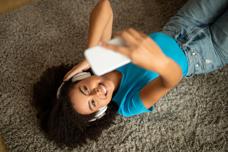 Smiling millennial african american curly lady in wireless headphones listen to music, lies on floor with smartphoneの写真素材