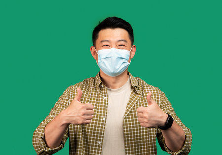 Asian middle aged man wearing face mask and gesturing thumbs up, approving personal protection on green backgroundの写真素材
