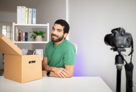 Happy attractive millennial arab guy with beard at workplace with camera and cardboard boxの写真素材