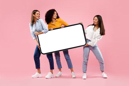 Excited diverse female friends holding huge smartphone with empty screen on pink studio background, mockupの写真素材
