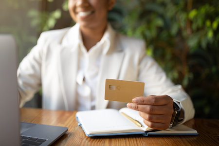 Glad millennial african american businesswoman in white suit uses laptop and credit card in cafe