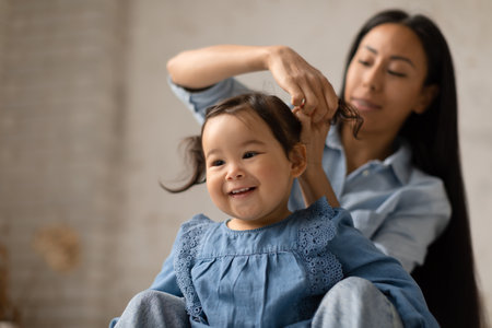 Cheerful Japanese Mommy Doing Baby Daughters Hair Making Ponytails Indoorの写真素材