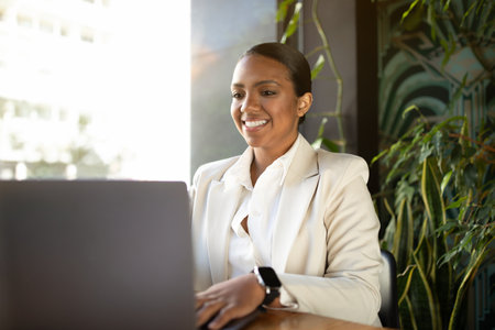 Positive millennial african american businesswoman in white suit working on laptop in cafeの写真素材
