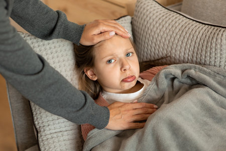 Worry millennial woman touching forehead of sad teen european girl, suffering from illness, lies on sofaの写真素材
