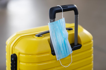 Safe Travel During Pandemic. Protective Medical Face Mask Hanged On Yellow Suitcaseの写真素材