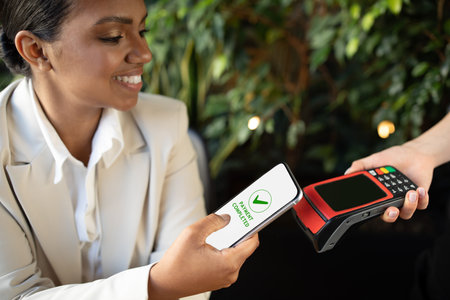 Happy millennial african american woman in white suit pays money bill by smartphone to terminal in...