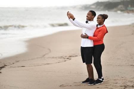 Smiling millennial african american family in sportswear in headphones take selfie on beach near oceanの写真素材