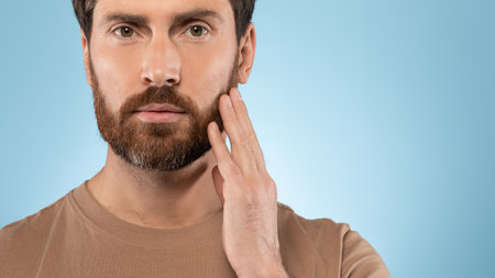 Headshot portrait of middle aged man touching beard and looking at camera over blue background, panorama, free spaceの写真素材