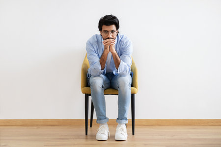 Pensive young middle eastern male in casual and glasses sits on armchair think, create idea in room