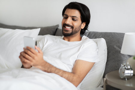 Happy young middle eastern guy in white t-shirt typing on smartphone, lies on bed, enjoy free time