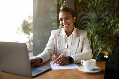 Smiling millennial african american businesswoman, boss in white suit working on laptop, making notesの写真素材