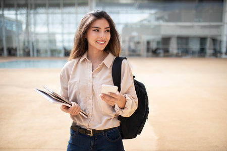 Glad millennial mixed race female student with backpack, books and smartphone in university campus, outdoorの写真素材
