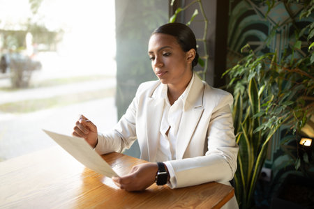 Serious concentrated millennial african american businesswoman in white suit reading document in cafeの写真素材