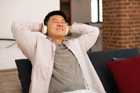 Happy asian middle aged man listening music in headphones, leaning on sofa with closed eyes and hands behind headの写真素材