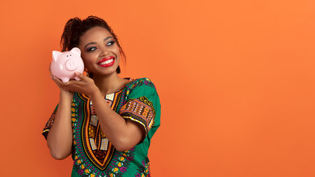 Cheerful african woman holding piggy bank next to earの写真素材