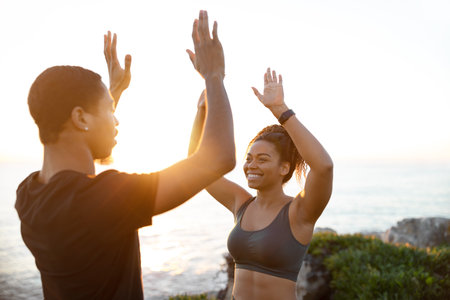 Glad millennial african american couple in sportswear give high five on ocean beach, sun flareの写真素材