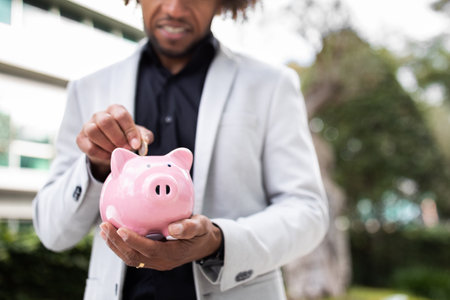 Savings, investment, business profit. Unrecognizable black businessman putting money in piggy bank outdoors, croppedの写真素材