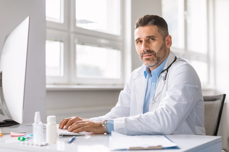 Online consultation service concept. Confident middle aged male GP doctor using computer, sitting at desk in officeの写真素材