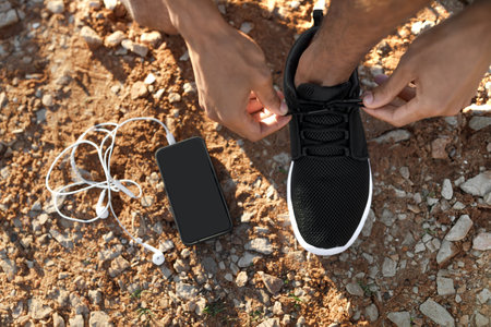 Millennial african american man tying shoelaces on sneakers on road or beach with headphones, phone