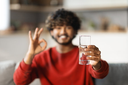 Smiling Young Indian Guy Holding Glass Of Water And Showing Ok Gestureの写真素材