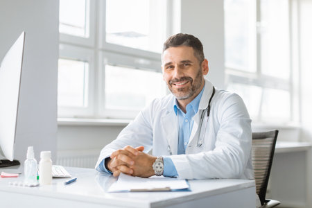 Portrait of happy cheerful man in white workwear doctor posing at clinic, sitting at workdesk and smiling at cameraの写真素材