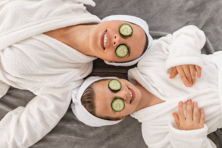 Happy mother and daughter in bathrobes lying with cucumber slices on eyesの写真素材