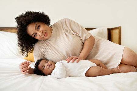 Parenthood and toddler. Glad pretty young african american lady and little child lie on bed, relax togetherの写真素材
