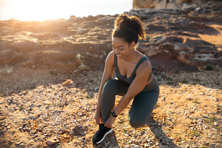 Positive millennial african american female in sportswear tying shoelaces on sneakers, on ocean...