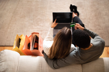 Millennial arabic husband hug caucasian wife with packages with purchase uses laptop with empty screenの写真素材