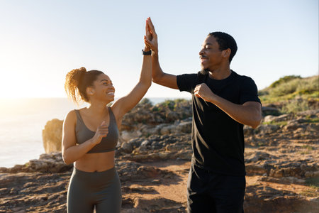 Positive millennial african american husband and wife in sportswear running, give high five on ocean beachの写真素材