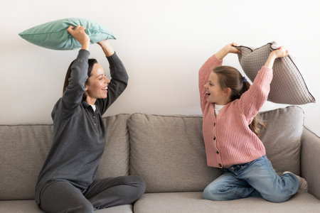 Cheerful excited european millennial mother and little girl fight pillow, have fun together in living roomの写真素材