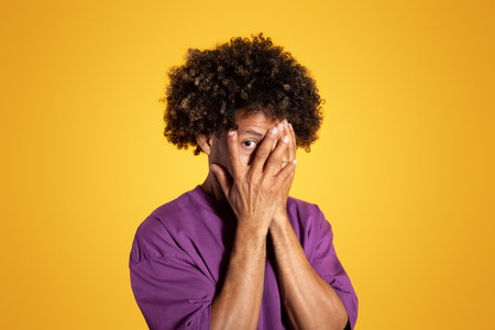Sad mature african american curly man in purple t-shirt, covers face with hands on yellow studio backgroundの写真素材