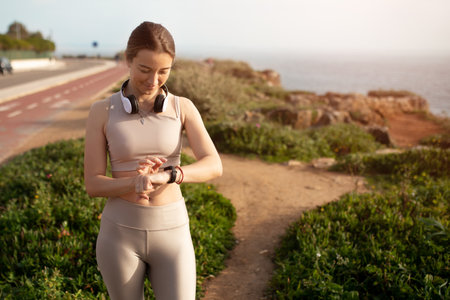 Smiling young caucasian lady in sportswear, wireless headphones, checks fitness tracker, control...