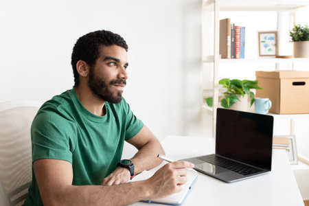 Smiling young middle eastern guy blogger or manager at table with laptop with blank screenの写真素材