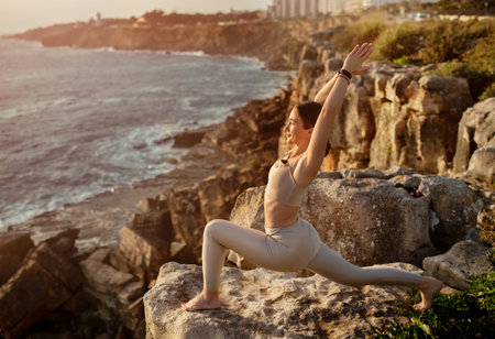 Satisfied young caucasian lady in sportswear practicing yoga on rocks, enjoy ocean, sunrise and training outdoorの写真素材