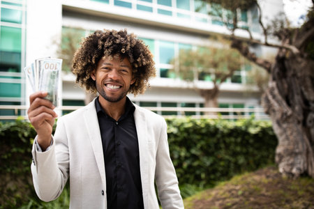 Happy rich black middle aged businessman holding money cash, celebrating financial luck and success, standing outdoorsの写真素材