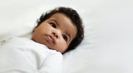 Portrait of cute curly african american small newborn baby lies on soft white bed in bedroom...