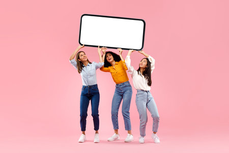 Cellphone display template. Three diverse ladies holding huge smartphone with blank screen over pink background, mockupの写真素材