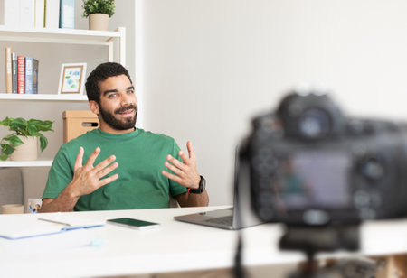 Glad young arabic man blogger at table with laptop working remotely, shooting vlog, online lessonの写真素材