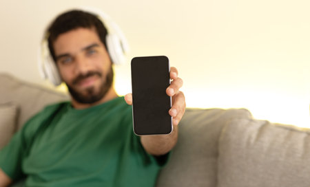 Glad young middle eastern man in wireless headphones shows smartphone with blank screen, enjoys musicの写真素材